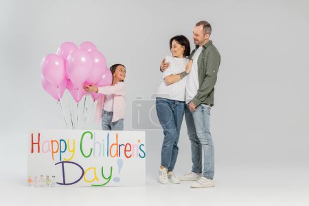 Longitud completa de padres sonrientes con ropa casual abrazando y mirando a la hija preadolescente parada cerca de globos rosados y pancarta con letras felices del día de los niños sobre fondo gris
