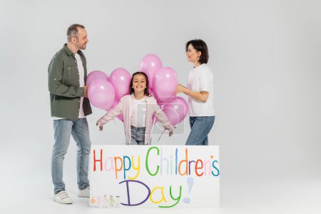 Smiling parents in casual clothes looking at each other near preteen daughter, pink festive balloons and placard with happy children's day lettering on grey background