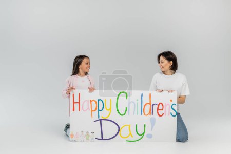 Smiling preteen girl looking at brunette and positive mother near placard with happy children's day lettering during celebration in June on grey background with copy space 