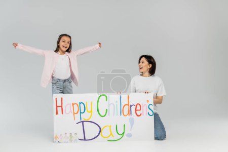 Excited preteen girl in casual clothes looking away while standing near mother and placard with happy children's day lettering during celebration on grey background