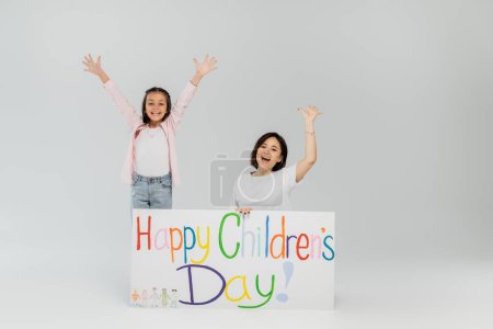 Excited preteen girl in casual clothes waving hands at camera near mother and placard with happy children's day lettering during celebration in June on grey background