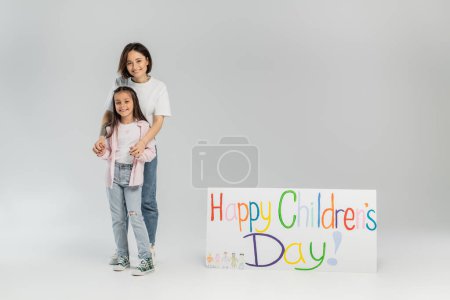 Adult woman in casual clothes hugging cheerful preteen daughter and looking at camera near placard with happy children's day lettering during celebration on grey background