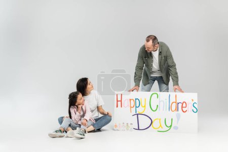 Lächelnde Frau und schwangere Tochter schauen Vater an, der neben Plakat mit fröhlichem Kindertagsaufdruck bei Feier im Juni auf grauem Hintergrund mit Kopierplatz steht