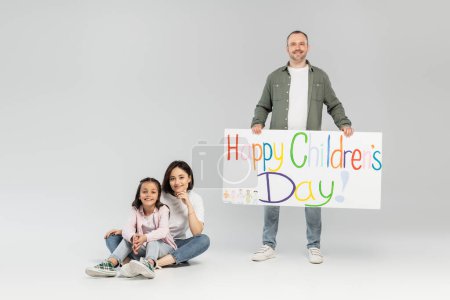 Full length of smiling father holding placard with happy children's day lettering near wife and preteen daughter looking at camera while sitting and celebrating in June on grey background