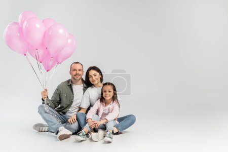 Sonrientes padres con ropa casual abrazando a su hija preadolescente y sosteniendo globos rosados festivos mientras celebran el día internacional de los niños sobre un fondo gris