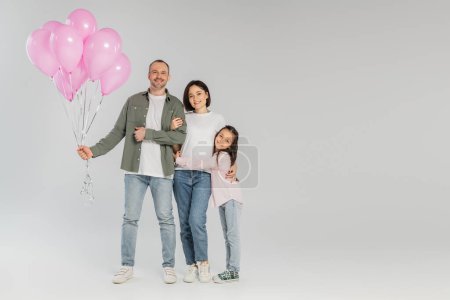 Full length of cheerful man holding festive pink balloons near wife and preteen daughter while celebrating child protection day and looking at camera on grey background