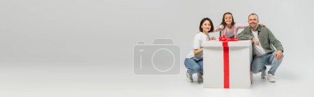 Carefree preteen girl in casual clothes hugging parents and looking at camera near big present with bow during child protection day celebration on grey background with copy space, banner 