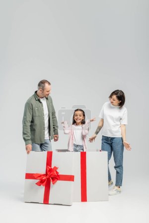 Volle Länge fröhlicher Eltern, die beim Kinderschutztag auf grauem Hintergrund in die Kamera schauen, während sie in einem großen Geschenkkarton stehen