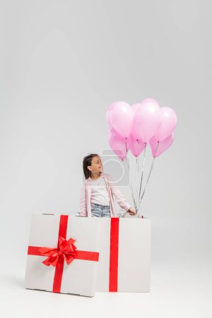 Smiling preteen kid in casual clothes looking at pink balloons while standing in big gift box during happy children day celebration on grey background