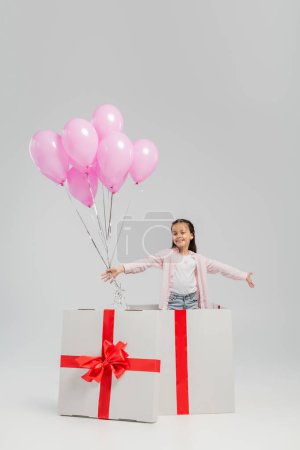 Niño preadolescente despreocupado con ropa casual mirando a la cámara mientras sostiene globos festivos rosados mientras está de pie en un gran regalo durante la celebración del día internacional de los niños sobre un fondo gris