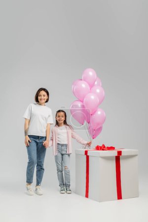 Photo for Full length of smiling tattooed mother holding hand of preteen daughter near pink balloons and big gift box during children day celebration on grey background - Royalty Free Image
