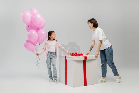 Full length of smiling preteen girl holding pink balloons and looking at tattooed mom near big present box during happy children day celebration on grey background