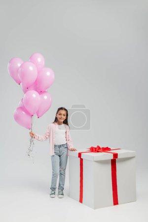 Longitud completa de niño preadolescente despreocupado en ropa casual mirando a la cámara mientras sostiene globos rosados cerca de regalo grande durante el día de protección infantil sobre fondo gris
