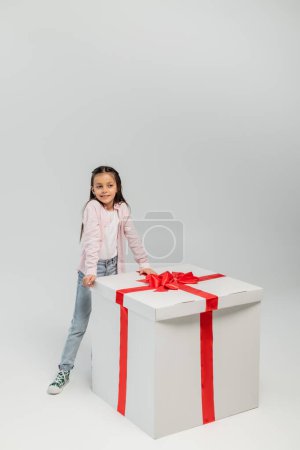 Longitud completa de niño preadolescente sonriente con ropa casual de pie cerca de un gran regalo con lazo mientras celebra el día internacional de los niños sobre un fondo gris