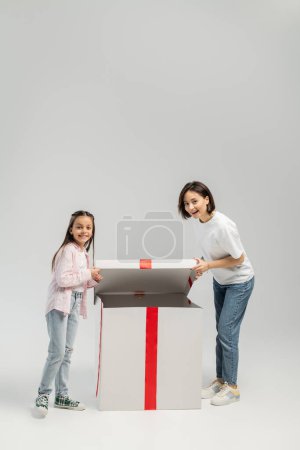 Volle Länge der fröhlichen Mutter und Tochter in lässiger Kleidung, die in die Kamera schaut, während sie während des internationalen Kindertages eine große Geschenkschachtel auf grauem Hintergrund öffnet