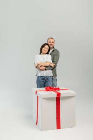 Smiling man in casual clothes hugging wife and looking at camera near big present with bow during international children day celebration on grey background
