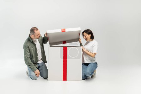 Excited tattooed woman looking at open big present box near husband during child protection day celebration on grey background with copy space 