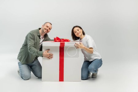 Lächelndes erwachsenes Paar in lässiger Kleidung, das in die Kamera schaut, während es die große Geschenkbox umarmt und beim Kinderschutztag auf grauem Hintergrund in die Kamera schaut