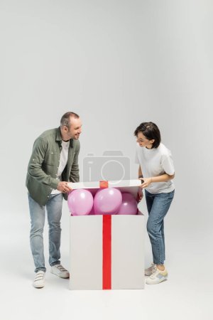 Full length of smiling adult couple in casual clothes opening big gift box with pink balloons during child protection day celebration while standing on grey background