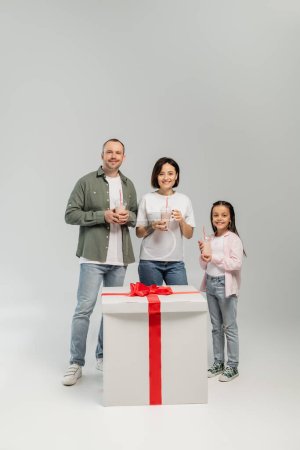 Positive Familie mit frühgeborener Tochter, die Milchshakes in die Kamera hält und in der Nähe einer großen Geschenkbox während der Kinderschutztagsfeier auf grauem Hintergrund blickt