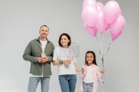 Lächelnde Eltern und ihre schwangere Tochter halten rosa Luftballons und Milchshakes in die Kamera, während sie während der internationalen Kindertagsfeier isoliert auf grau schauen 