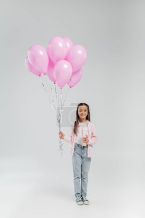 Longitud completa de la niña preadolescente sonriente en ropa casual sosteniendo globos rosados y batido mientras mira a la cámara durante la celebración del día de protección infantil sobre fondo gris