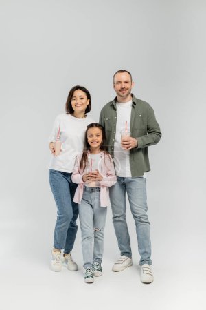 Full length of smiling family with preteen daughter in casual clothes holding milkshakes in plastic cups and looking at camera during children day celebration on grey background