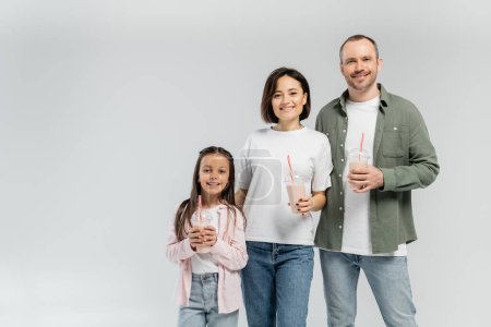 Positive parents in casual clothes looking at camera near daughter with milkshake in plastic cup during international children day celebration isolated on grey with copy space