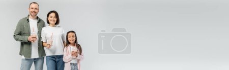 Photo for Smiling adult parents in casual clothes holding milkshakes near preteen kid and looking at camera during international child protection day isolated on grey with copy space, banner - Royalty Free Image