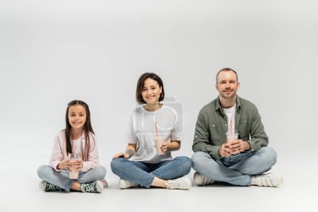 Smiling adult parents in casual clothes looking at camera while holding plastic cups with milkshakes near preteen daughter during international children day celebration and sitting on grey background