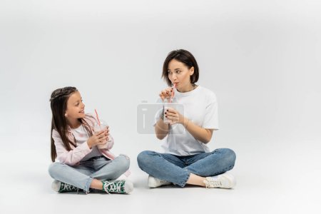 Fröhliches Mädchen in lässiger Kleidung hält Plastikbecher mit Milchshake mit Trinkhalmen in der Hand und blickt tätowierte Mutter an, die während der Feier zum Internationalen Kinderschutztag auf grauem Hintergrund sitzt