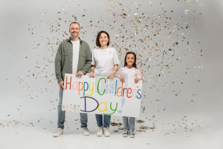 Full length of cheerful family with preteen daughter in casual clothes holding placard with happy children's day lettering and standing under falling festive confetti on grey background