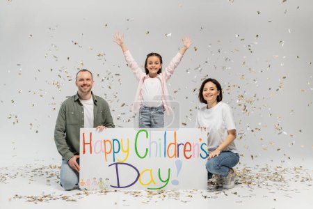 Excited preteen girl in casual clothes waving hands at camera while standing near parent holding placard with happy children's day lettering and under falling festive confetti on grey background