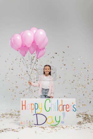 Fröhliches Frühchen in legerer Kleidung blickt in die Kamera und hält rosa Luftballons in der Nähe eines Plakats mit fröhlichem Kindertagsaufdruck und unter fallendem Festkonfetti auf grauem Hintergrund.