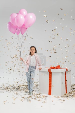Unbekümmertes Mädchen in lässiger Kleidung mit rosa Luftballons und Blick in die Kamera in der Nähe einer großen Geschenkschachtel, während es bei der Feier zum Kinderschutztag unter Konfetti auf grauem Hintergrund steht