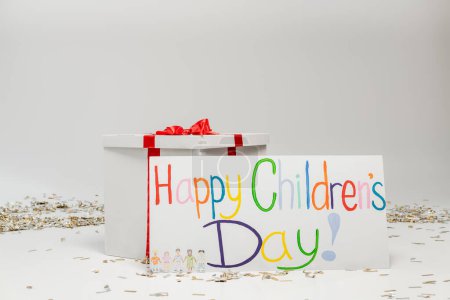 Placard con letras coloridas del día de los niños felices cerca de la caja de regalo grande con lazo en fondo gris con confeti festivo 
