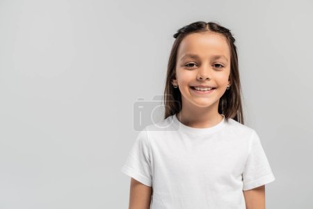Portrait of smiling preteen and brunette girl in white t-shirt looking at camera while celebrating global child protection day isolated on grey with copy space