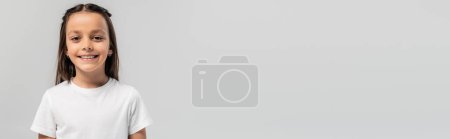 Téléchargez les photos : Portrait de préadolescente brune insouciante en t-shirt blanc regardant la caméra pendant la célébration de la Journée internationale de la protection de l'enfance isolé sur gris avec espace de copie, bannière - en image libre de droit