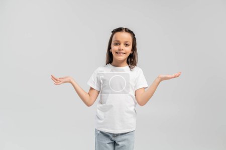 Smiling brunette preteen kid in white t-shirt and jeans looking at camera and showing shrug gesture while celebrating child protection day and standing isolated on grey 