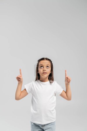 Focused preteen girl in white t-shirt and jeans pointing with fingers and looking up during international child protection day celebration isolated on grey with copy space 