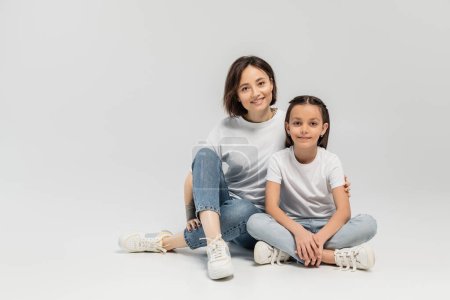 happy woman with tattoo on hand and short hair hugging preteen daughter while sitting together in white t-shirts and blue denim jeans on grey background, international children's day 