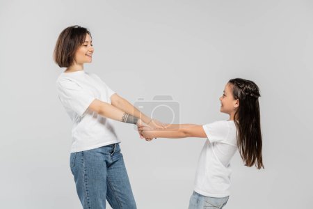 happy mother with tattoo on hand and short hair holding hands with joyous daughter while standing together in white t-shirts and blue denim jeans on grey background, child protection day