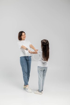 full length of happy mother with tattoo on hand and short hair holding hands with preteen daughter while standing together in white t-shirts and blue denim jeans on grey background 
