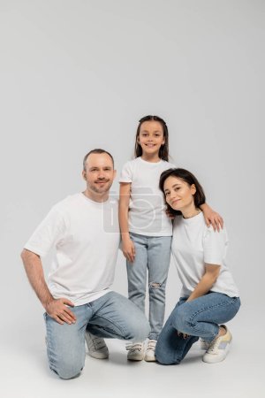 joyous father and mother with short hair sitting near cheerful preteen daughter while posing together in white t-shirts and blue denim jeans on grey background, Happy children's day