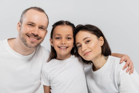 portrait of happy family in white t-shirts looking at camera on grey background, Child protection day, cheerful father and mother with short hair embracing preteen daughter 