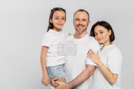 retrato de familia feliz en camisetas blancas mirando a la cámara sobre fondo gris, Día de la protección del niño, padre alegre levantando hija preadolescente cerca de la esposa con pelo corto y tatuaje 