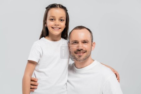 unshaved and cheerful father with bristle hugging happy preteen daughter with long brunette hair while posing in white t-shirts and looking at camera on grey background, Child protection day 