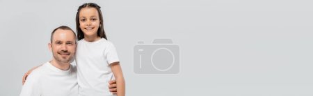 unshaved and cheerful father with bristle hugging happy preteen daughter with long brunette hair while posing in white t-shirts and looking at camera on grey background, Child`s protection day, banner
