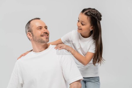 glückliches Mädchen mit langen brünetten Haaren, angelehnt an einen unrasierten und fröhlichen Vater mit Borsten, während sie in weißen T-Shirts posiert und sich auf grauem Hintergrund anschaut, Kinderschutztag 