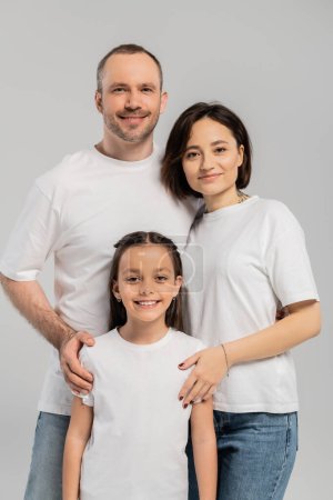 portrait of happy family in white t-shirts looking at camera on grey background, International child protection day, father and mother with tattoo embracing preteen brunette daughter 